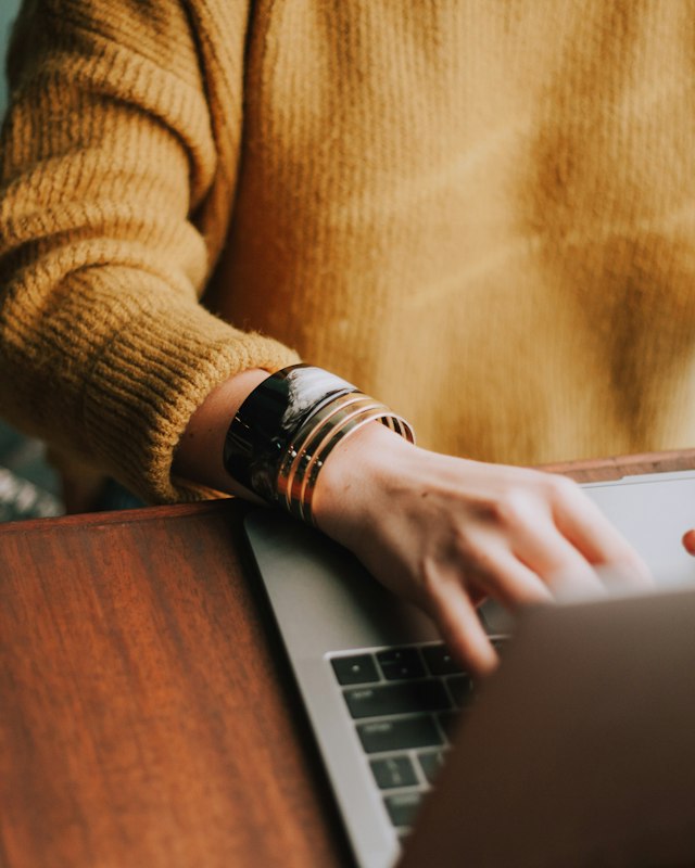 Person in a mustard sweater typing on a laptop at a wooden desk